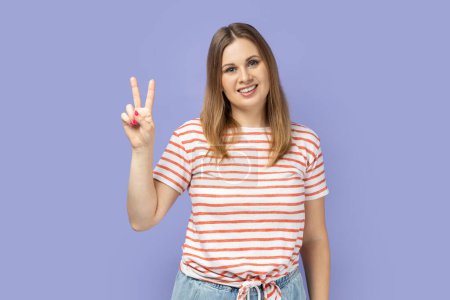 Photo for Portrait of beautiful blond woman wearing striped T-shirt smiling happily and showing v sign or peace gesture, rejoicing win. Indoor studio shot isolated on purple background. - Royalty Free Image