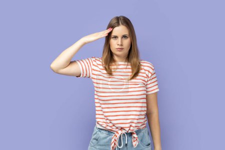 Responsible patriotic blond woman wearing striped T-shirt following discipline, saluting to commander with hand near temple and listening order. Indoor studio shot isolated on purple background.