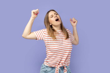 Photo for Portrait of tired blond woman wearing striped T-shirt standing yawning with closed eyes, stretching arms, feeling exhausted need rest. Indoor studio shot isolated on purple background. - Royalty Free Image