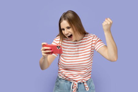 Photo for Portrait of excited smiling woman gamer wearing striped T-shirt playing game on smart phone, winning level, clenched fist, celebrating her victory. Indoor studio shot isolated on purple background. - Royalty Free Image