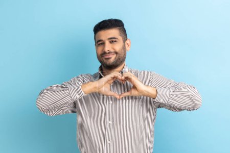 Photo for Happy positive handsome businessman with beard standing and showing heart shape making with hands, looking at camera with love, wearing striped shirt. Indoor studio shot isolated on blue background. - Royalty Free Image