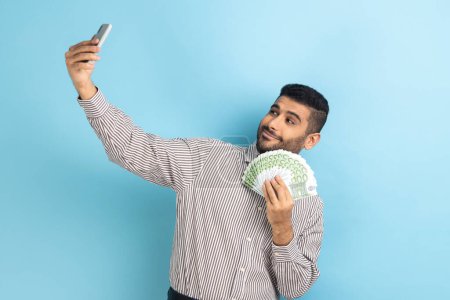 Photo for Rich businessman holding money and taking selfie by mobile phone, smiling boasting of big profit, successful online betting, wearing striped shirt. Indoor studio shot isolated on blue background. - Royalty Free Image