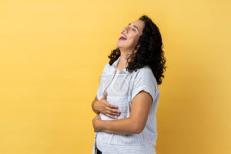 Photo for Portrait of overjoyed woman with dark wavy hair laughing happily at something keeps hands on belly, smiles broadly, expressing positive emotions.Indoor studio shot isolated on yellow background. - Royalty Free Image