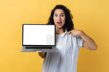 Photo for Portrait of amazed surprised woman with dark wavy hair holding and pointing laptop with empty display, copy space for advertisement. Indoor studio shot isolated on yellow background. - Royalty Free Image
