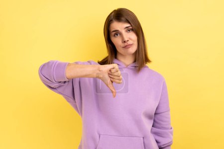 Photo for Portrait of upset dissatisfied woman standing with thumb down, negative feedback, demonstrating disapproval gesture, wearing purple hoodie. Indoor studio shot isolated on yellow background. - Royalty Free Image