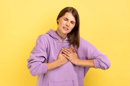 Photo for Portrait of overworked depressed woman frowning suffering sudden heart attack, myocardial infarction, risk of breast cancer, wearing purple hoodie. Indoor studio shot isolated on yellow background. - Royalty Free Image