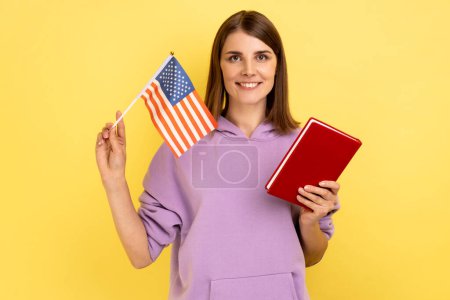 Foto de Retrato de una atractiva estudiante morena sosteniendo libro y bandera americana, educación en el extranjero en Estados Unidos, con capucha púrpura. Estudio interior plano aislado sobre fondo amarillo. - Imagen libre de derechos