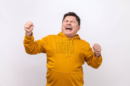 Photo for Overjoyed handsome middle aged man expressing winning gesture with raised fists and screaming, celebrating victory, wearing urban style hoodie. Indoor studio shot isolated on white background. - Royalty Free Image