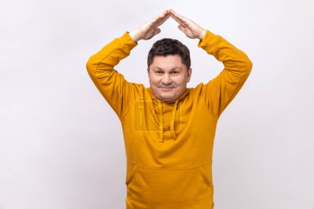 Photo for Portrait of cheerful middle aged man standing under roof house gesture and smiling, dreaming of real estate purchase, wearing urban style hoodie. Indoor studio shot isolated on white background. - Royalty Free Image