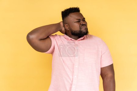 Photo for Portrait of worried anxious bearded man wearing pink shirt touching neck, feeling acute pain moving and turning head, suffering spine problems. Indoor studio shot isolated on yellow background. - Royalty Free Image