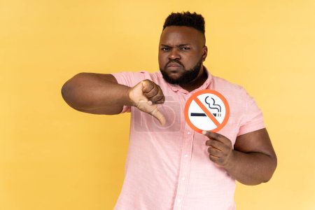 Photo for Portrait of serious young adult bearded man wearing pink shirt holding no smoking sign, showing thumb down gesture, looking at camera. Indoor studio shot isolated on yellow background. - Royalty Free Image