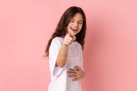 Photo for Portrait of positive optimistic joyful little girl wearing white T-shirt laughing out loud holding belly and pointing finger on you, mockery. Indoor studio shot isolated on pink background. - Royalty Free Image