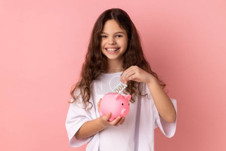 Photo for Portrait of delighted smiling little girl wearing white T-shirt standing looking at camera, putting dollar banknote into piggy bank. Indoor studio shot isolated on pink background. - Royalty Free Image
