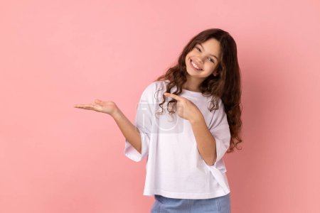 Photo for Portrait of cute little girl wearing white T-shirt presenting copy space on her palm, showing empty place for commercial text or goods. Indoor studio shot isolated on pink background. - Royalty Free Image