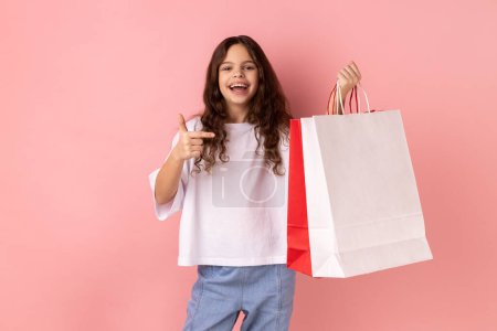 Foto de Retrato de una niña excitada con una camiseta blanca apuntando a las bolsas de la compra, mirando a la cámara con feliz expresión facial. Estudio interior plano aislado sobre fondo rosa. - Imagen libre de derechos