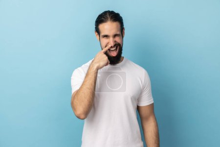 Photo for Portrait of crazy handsome man with beard wearing white T-shirt standing and drilling his nose with funny face, winking at camera. Indoor studio shot isolated on blue background. - Royalty Free Image