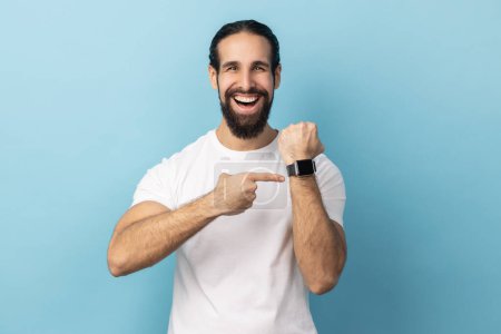 Téléchargez les photos : Look at clock, no rush. Portrait of man with beard wearing white T-shirt pointing wrist watch and expressing optimism about time, not busy. Indoor studio shot isolated on blue background. - en image libre de droit