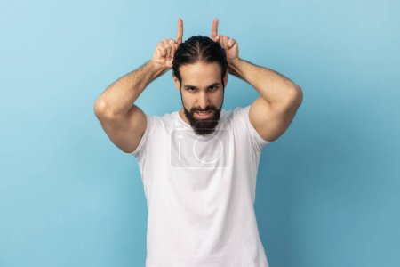 Foto de Portrait of aggressive bully man with beard wearing white T-shirt showing bull horns gesture over head, frowning as before attack, Indoor studio shot isolated on blue background. - Imagen libre de derechos