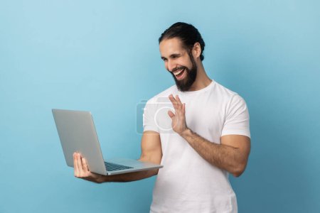 Photo for Portrait of smiling delighted man with beard wearing white T-shirt having video call, waving hand to camera, saying hello or good bye. Indoor studio shot isolated on blue background. - Royalty Free Image
