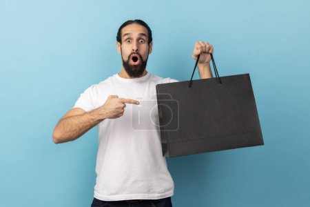 Photo for Portrait of shocked man with beard wearing white T-shirt pointing at shopping bags, looking at camera with astonished surprised facial expression. Indoor studio shot isolated on blue background. - Royalty Free Image