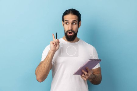 Photo for Portrait of man with beard wearing white T-shirt writing creative idea into notebook, taking notes in paper, raised arm, keeps mouth open. Indoor studio shot isolated on blue background. - Royalty Free Image