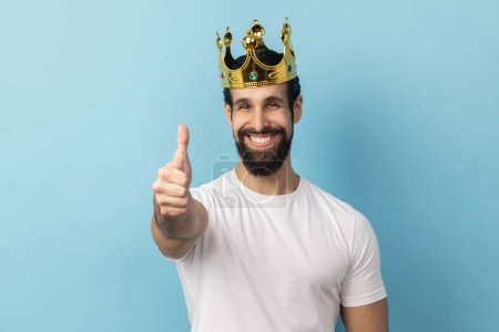 Foto de Portrait of smiling joyful man with beard wearing white T-shirt and in gold crown standing looking at camera with toothy smile, showing thumb up. Indoor studio shot isolated on blue background. - Imagen libre de derechos
