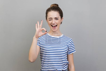Foto de Portrait of funny playful positive young adult woman wearing striped T-shirt winking to camera and showing ok sign, approval gesture. Indoor studio shot isolated on gray background. - Imagen libre de derechos