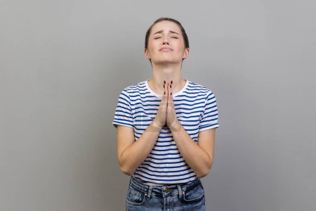 Téléchargez les photos : Please, I'm begging. Portrait of upset worried woman wearing striped T-shirt looking up with imploring desperate grimace, praying to god asking for help. Indoor studio shot isolated on gray background - en image libre de droit