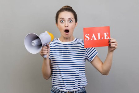 Foto de Portrait of shocked astonished amazed woman wearing striped T-shirt holding card with sale inscription and megaphone, making announcement. Indoor studio shot isolated on gray background. - Imagen libre de derechos