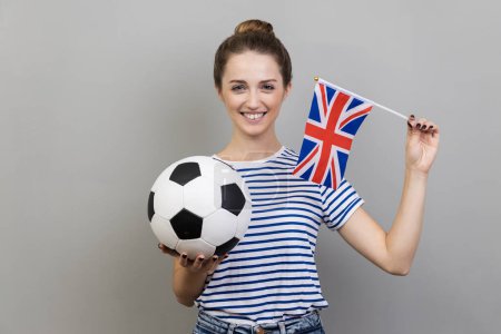 Téléchargez les photos : Portrait of woman wearing striped T-shirt holding british flag and black and white ball, cheering for his favorite team, supporting. Indoor studio shot isolated on gray background. - en image libre de droit