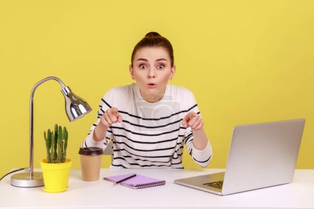 Foto de Extremely shocked woman office worker in striped shirt sitting workplace, looking and pointing at camera with astonished surprised face. Indoor studio studio shot isolated on yellow background. - Imagen libre de derechos