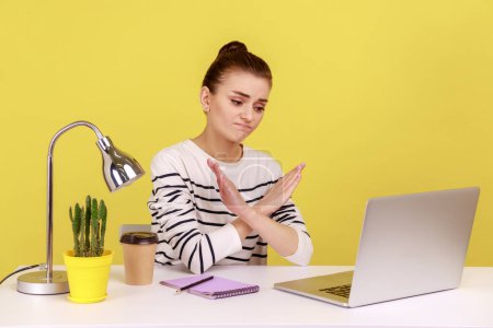 Téléchargez les photos : Upset sad woman manager sitting at workplace crossing hands showing x sign, looking at laptop screen with frowning face. Indoor studio studio shot isolated on yellow background. - en image libre de droit