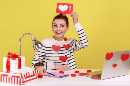 Téléchargez les photos : Woman covered with heart sticks holding likes counter template above head sitting at workplace with laptop, making posts in social networks. Indoor studio studio shot isolated on yellow background. - en image libre de droit