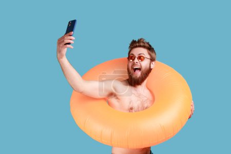 Foto de Portrait of excited bearded man posing with orange rubber ring and holding mobile phone, making selfie, enjoying his vacation at seaside. Indoor studio shot isolated on blue background. - Imagen libre de derechos
