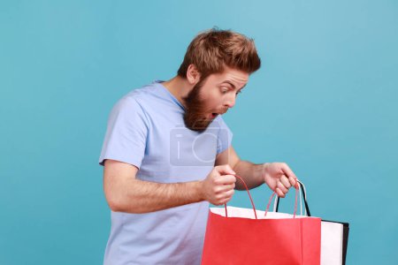 Photo pour Portrait of attractive bearded man standing, holding red and white shopping bags and looking inside with shocked facial expression. Indoor studio shot isolated on blue background. - image libre de droit