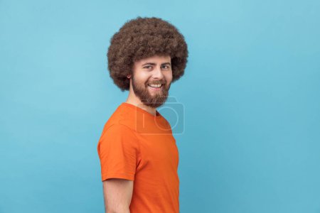 Foto de Side view of man wearing orange T-shirt looking at camera with toothy smile and happy facial expression, being in good mood, rejoicing great results. Indoor studio shot isolated on blue background. - Imagen libre de derechos