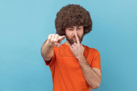 Foto de Portrait of man with Afro hairstyle wearing orange T-shirt touching nose, showing liar gesture, angry about falsehood, pointing to camera fake news. Indoor studio shot isolated on blue background. - Imagen libre de derechos