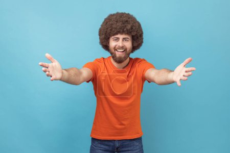 Téléchargez les photos : Come into my arms. Portrait of satisfied man with Afro hairstyle wearing orange T-shirt reaching out to camera, stretching arms to hug you. Indoor studio shot isolated on blue background. - en image libre de droit