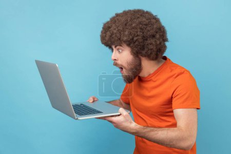 Foto de Man with Afro hairstyle in orange T-shirt doing freelance job on laptop, typing email or surfing internet, looking surprised at laptop screen. Indoor studio shot isolated on blue background. - Imagen libre de derechos