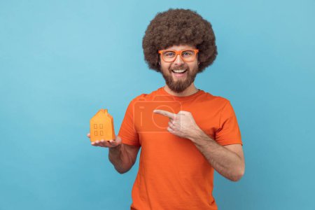 Téléchargez les photos : Portrait of smiling man with Afro hairstyle wearing orange T-shirt and funny eyeglasses holding pointing at paper house, helping with rent. Indoor studio shot isolated on blue background. - en image libre de droit