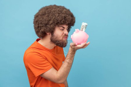 Photo for Portrait of crazy angry man with Afro hairstyle wearing orange T-shirt standing holding looking at piggybank with dollar banknotes with frowning face. Indoor studio shot isolated on blue background. - Royalty Free Image