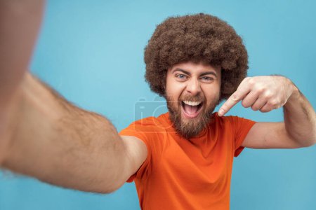 Foto de Portrait of smiling satisfied man blogger with Afro hairstyle wearing orange T-shirt broadcasting livestream, pointing down, asking to subscribe, POV. Indoor studio shot isolated on blue background. - Imagen libre de derechos