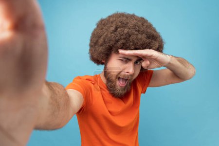 Foto de Portrait of positive handsome man with Afro hairstyle wearing orange T-shirt taking selfie for social networks, posing as he is looking far, POV. Indoor studio shot isolated on blue background. - Imagen libre de derechos