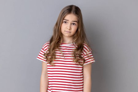 Téléchargez les photos : Portrait of unhappy little girl wearing striped T-shirt being upset of bad news, looking at camera with frowning face, expressing sadness. Indoor studio shot isolated on gray background. - en image libre de droit