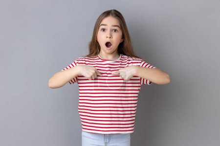 Foto de Portrait of astonished little girl wearing striped T-shirt pointing at herself, asks who me, has surprised expression, shocked being picked. Indoor studio shot isolated on gray background. - Imagen libre de derechos