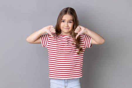 Photo for Portrait of unhappy sad little dark haired girl wearing striped T-shirt showing thumb down, expressing bad negative emotions, dislike. Indoor studio shot isolated on gray background. - Royalty Free Image