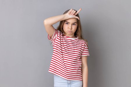 Photo for Portrait of depressed little girl wearing striped T-shirt showing loser gesture, L finger sign on forehead, upset about dismissal, unlucky day. Indoor studio shot isolated on gray background. - Royalty Free Image