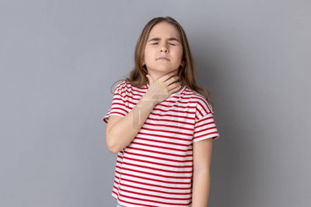 Téléchargez les photos : Portrait of little girl wearing striped T-shirt touching neck, suffering thyroid disorder or tonsillitis, inflamed throat, medical concept. Indoor studio shot isolated on gray background. - en image libre de droit