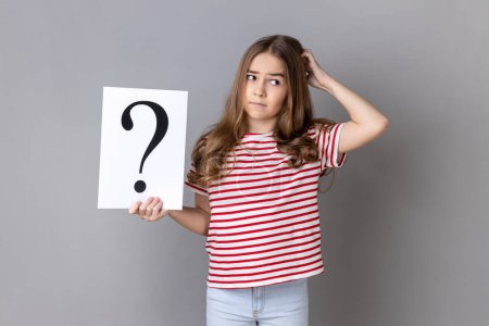 Portrait of little girl wearing striped T-shirt holding paper with question mark over, thoughtful, face thinking about question, very confused idea. Indoor studio shot isolated on gray background.