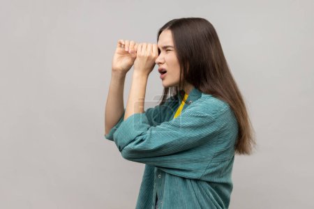 Foto de Side view of shocked young woman standing with hands on eye monocular gesture and looking away with surprised face, wearing casual style jacket. Indoor studio shot isolated on gray background. - Imagen libre de derechos